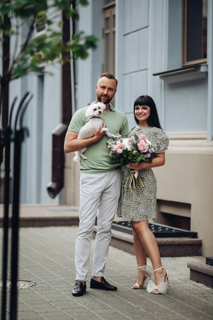 Feliz pareja romántica amorosa caminando con perro pequeño al aire libre. Chica con ramo de flores y chico con cachorro en citas