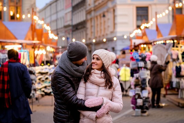 Feliz pareja romántica con ropa de invierno abrazándose mientras está de pie en la calle de la noche con la feria de Navidad