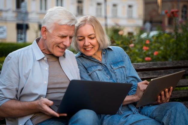 Feliz pareja senior al aire libre en un banco con ordenador portátil y tableta