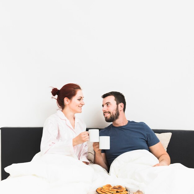 Feliz pareja sentada en la cama bebiendo café