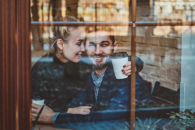 Una feliz pareja sonriente está sentada en un café y mirando por la ventana mientras disfruta de su café.