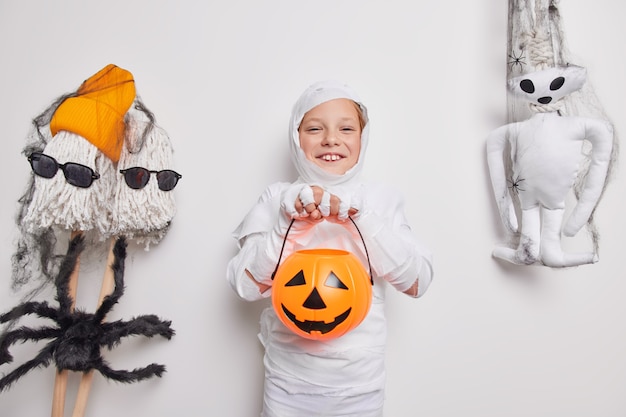 Feliz pequeño niño de halloween juega truco o trato calabaza jack o lantern envuelta en tela blanca rodeada de atributos de vacaciones aislados en blanco