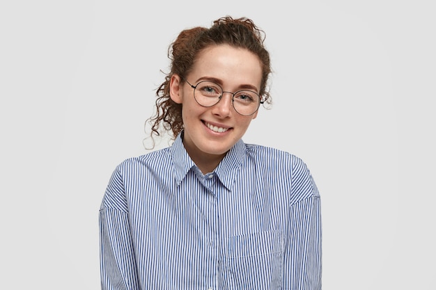 Foto gratuita feliz periodista feliz de tener una conversación agradable con un extraño, tiene una sonrisa suave, dientes blancos perfectos, piel pecosa, vestida con camisa a rayas, aislada en una pared blanca en blanco. concepto de positividad