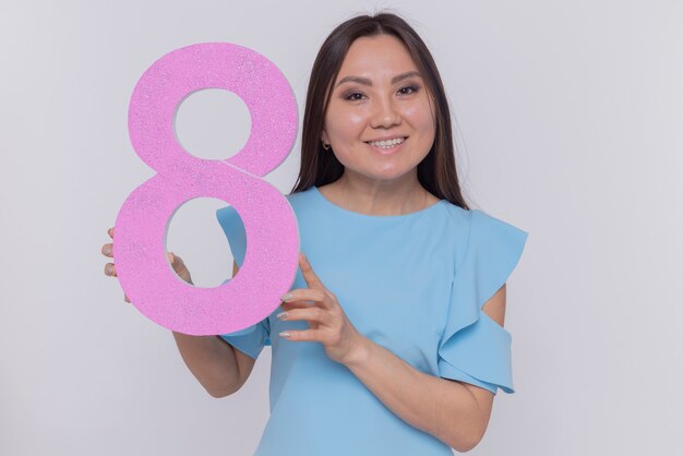 Feliz y positiva mujer asiática sosteniendo el número ocho de cartón mirando al frente sonriendo alegremente celebrando el día internacional de la mujer de pie sobre una pared blanca