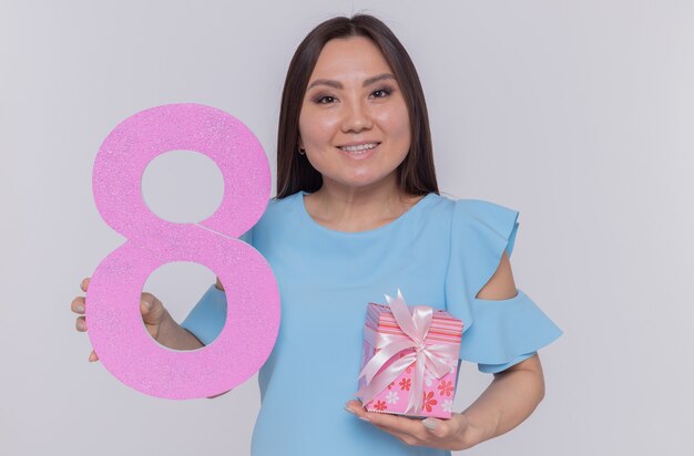 Feliz y positiva mujer asiática sosteniendo el número ocho hecho de cartón y presente mirando al frente sonriendo celebrando alegremente el día internacional de la mujer de pie sobre una pared blanca