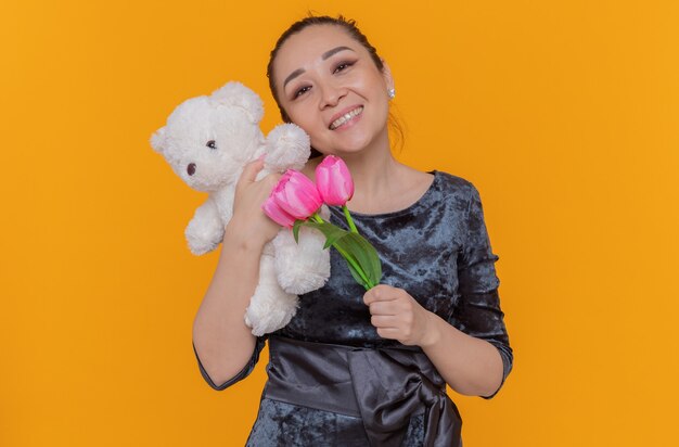 Feliz y positiva mujer asiática sosteniendo un ramo de tulipanes rosados y un oso de peluche sonriendo alegremente celebrando el día internacional de la mujer de pie sobre una pared naranja