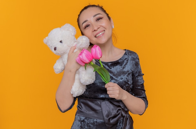 Feliz y positiva mujer asiática sosteniendo un ramo de tulipanes rosados y un oso de peluche sonriendo alegremente celebrando el día internacional de la mujer de pie sobre una pared naranja