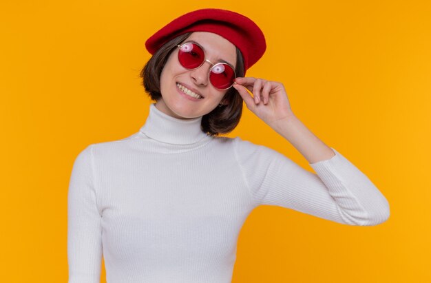 Feliz y positiva mujer joven con pelo corto en cuello alto blanco con boina y gafas de sol rojas mirando al frente sonriendo alegremente de pie sobre la pared naranja