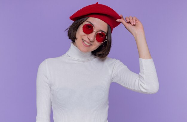 Feliz y positiva mujer joven con pelo corto en cuello alto blanco con boina y gafas de sol rojas mirando al frente sonriendo alegremente tocando su boina de pie sobre la pared azul