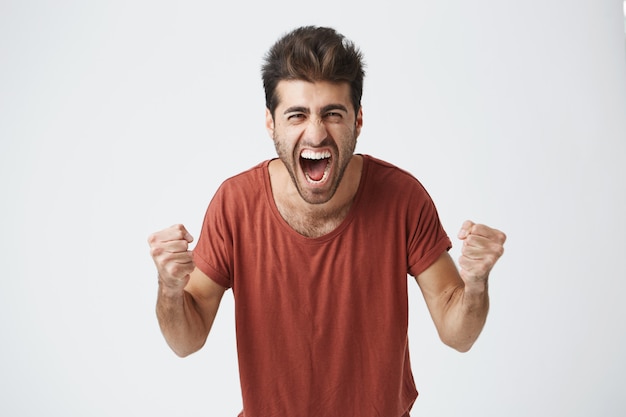 Foto gratuita feliz positivo emocionado joven apretando los puños y gritando, vistiendo una camiseta casual contento de escuchar buenas noticias, celebrando su victoria o éxito. logro de la vida, objetivos y concepto de felicidad