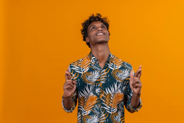Feliz y positivo joven apuesto hombre de piel oscura con cabello rizado en hojas camisa estampada sosteniendo los dedos juntos mientras mira hacia arriba sobre un fondo naranja