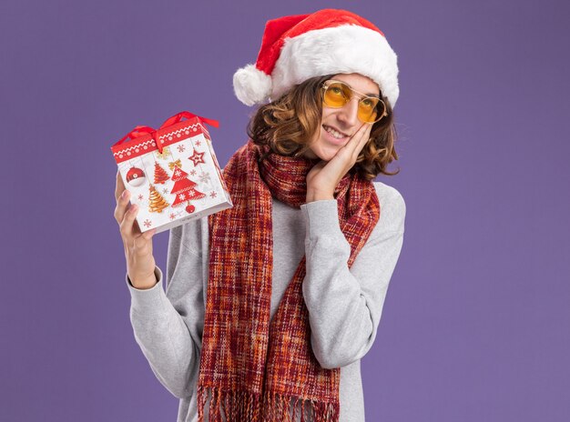 Feliz y positivo joven vestido con gorro de Papá Noel de Navidad y gafas amarillas con bufanda alrededor de su cuello sosteniendo un regalo de Navidad mirando hacia arriba sonriendo alegremente de pie sobre la pared púrpura