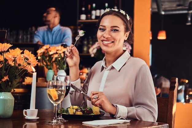 Feliz primer plano retrato de una hermosa mujer de piel negra con una blusa y una diadema de flores, disfrutando de la cena mientras come en un restaurante.