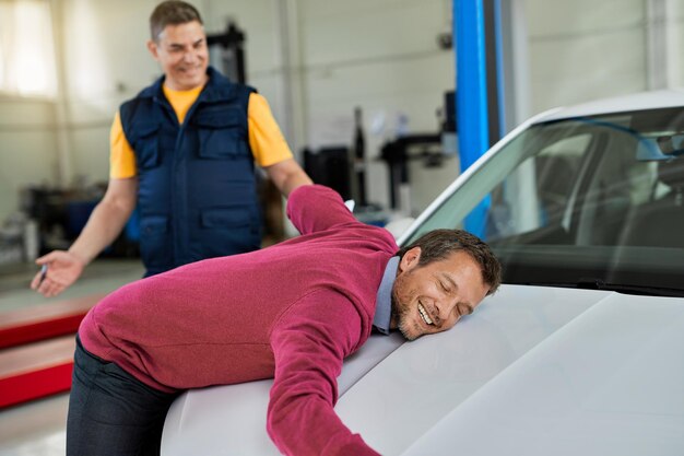 Feliz propietario de un automóvil sintiéndose aliviado y apoyado en el capó de un vehículo en un taller de reparación de automóviles