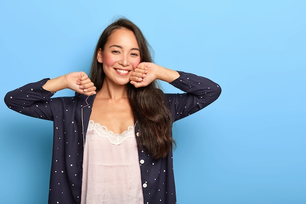 Feliz relajada joven asiática se despierta de buen humor, estira las manos durante la mañana, vestida con ropa de dormir