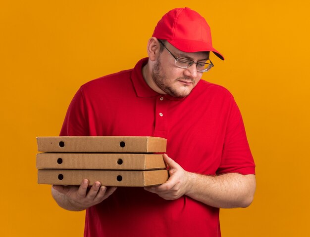 Feliz repartidor joven con sobrepeso en gafas ópticas sosteniendo cajas de pizza y mirando hacia abajo aislado en la pared naranja con espacio de copia