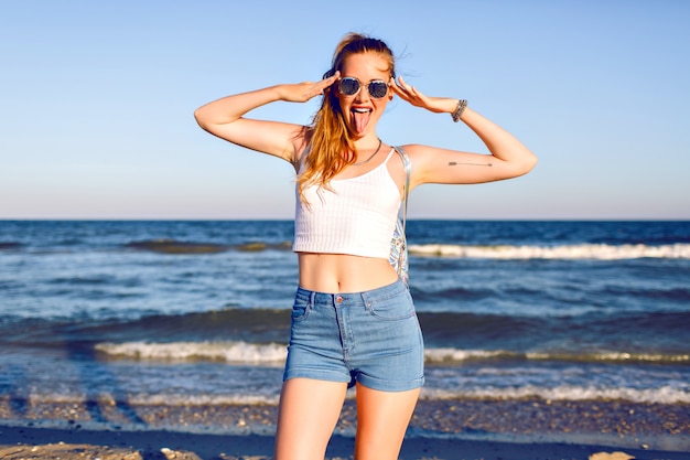 Foto gratuita feliz retrato positivo de joven rubia divirtiéndose en vacaciones tropicales, caminando cerca del océano, viaje con mochila, traje de verano con estilo hipster y gafas de sol. cuerpo sexy bronceado, pelos largos.