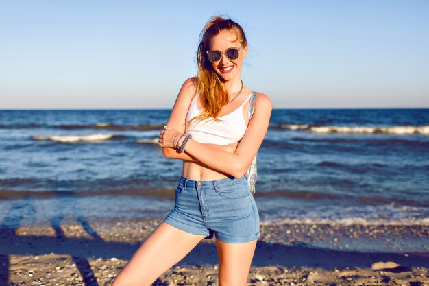 Feliz retrato positivo de joven rubia divirtiéndose en vacaciones tropicales, caminando cerca del océano, viaje con mochila, traje de verano con estilo hipster y gafas de sol. cuerpo sexy bronceado, pelos largos.