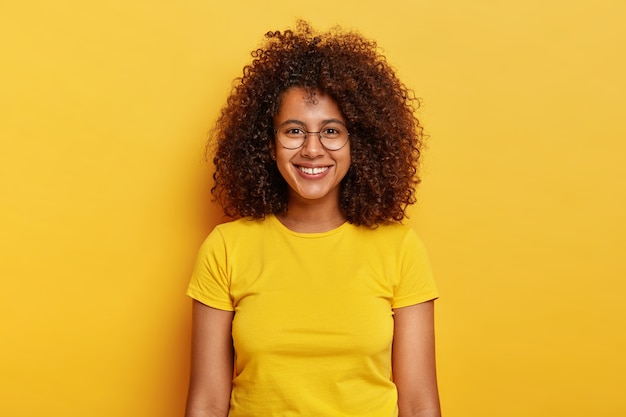 Feliz y seductora mujer joven con cabello oscuro y rizado, espera un evento emocionante, sonríe con alegría, usa grandes gafas redondas y una camiseta amarilla