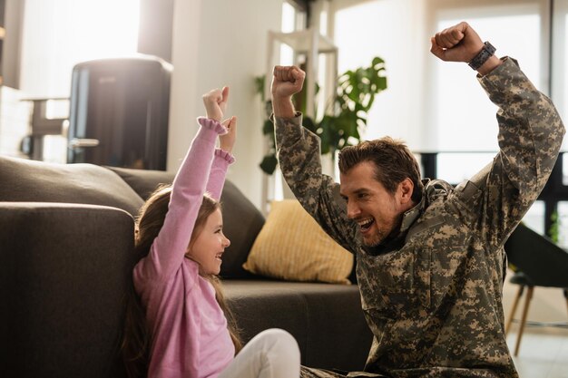 Feliz soldado del ejército y su pequeña hija divirtiéndose mientras celebran la reunión con los brazos levantados en la sala de estar