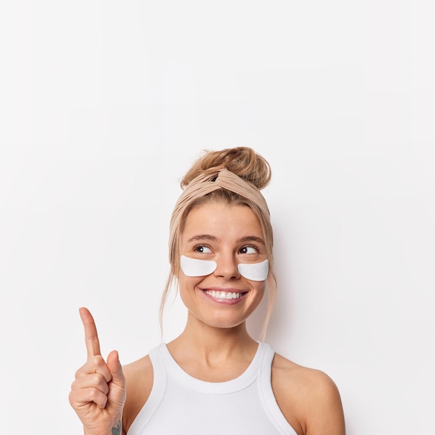 Feliz y soñadora mujer europea aplica parches de belleza debajo de los ojos para los puntos de cuidado de la piel con el dedo índice sobre la cabeza vestida con una camiseta informal aislada sobre fondo blanco cuida su apariencia