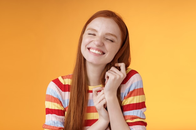 Feliz soñadora romántica joven tierna jengibre fantaseando creando la imaginación de la historia de amor sonriendo ampliamente encantada cerrar los ojos tocando mechones de cabello recordando un bonito recuerdo, de pie sobre fondo naranja.