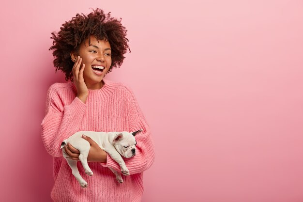 Feliz sonriente alegre hembra de piel oscura lleva un pequeño cachorro de bulldog francés dormido, viste un jersey rosa, enfocado a un lado, estando en alto espíritu, aislado sobre una pared rosa. Monocromo. Concepto de mascotas