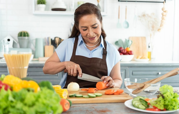 Feliz sonriente anciana asiática en delantal corta verduras mientras cocina ensalada en la cocina