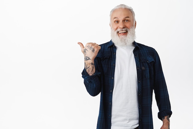 Feliz sonriente anciano abuelo barbudo con tatuajes apuntando a la izquierda en el texto promocional que muestra un anuncio con la cara emocionada de pie contra el fondo blanco