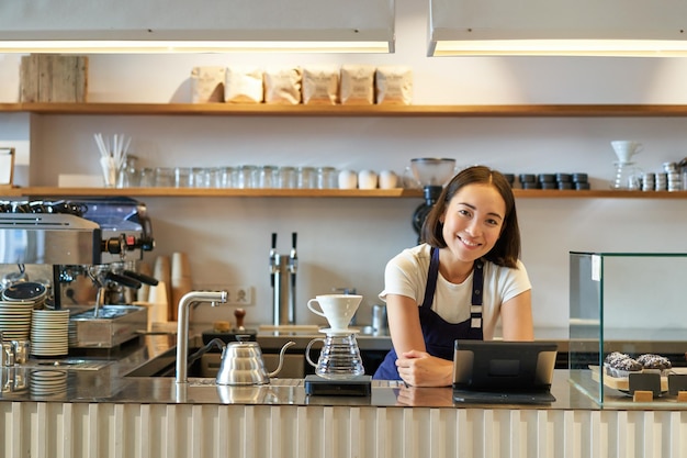 Feliz sonriente chica barista asiática detrás del mostrador trabajando con terminal pos y kit de filtro de elaboración de cerveza mak