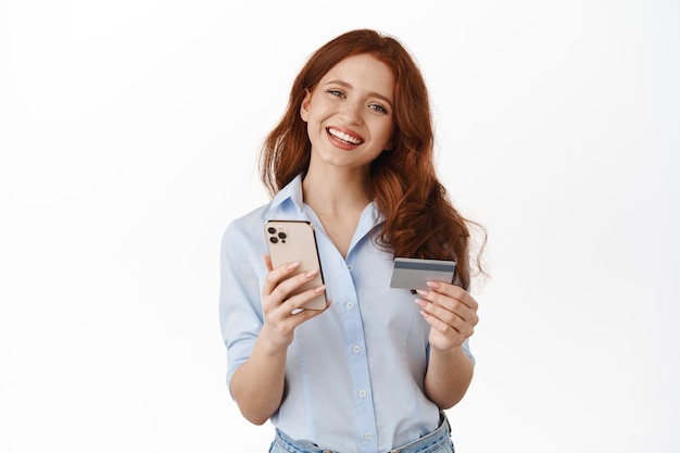 Feliz sonriente chica de compras sosteniendo teléfono móvil, tarjeta de crédito, pagando en la aplicación de teléfono inteligente, ordenando algo en el sitio, revisando la cuenta bancaria, de pie contra el fondo blanco.