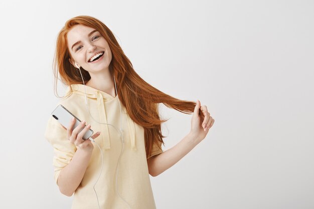 Feliz sonriente chica pelirroja escuchando música en auriculares y sosteniendo el teléfono móvil