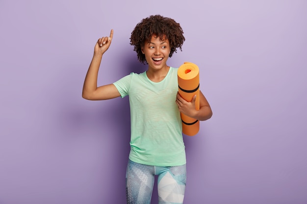 Foto gratuita feliz sonriente despreocupada deportista afroamericana de pelo rizado lleva estera de yoga arrugada, levanta el brazo e indica hacia arriba, disfruta de un buen entrenamiento, vestida con camiseta y leggings. concepto de deporte