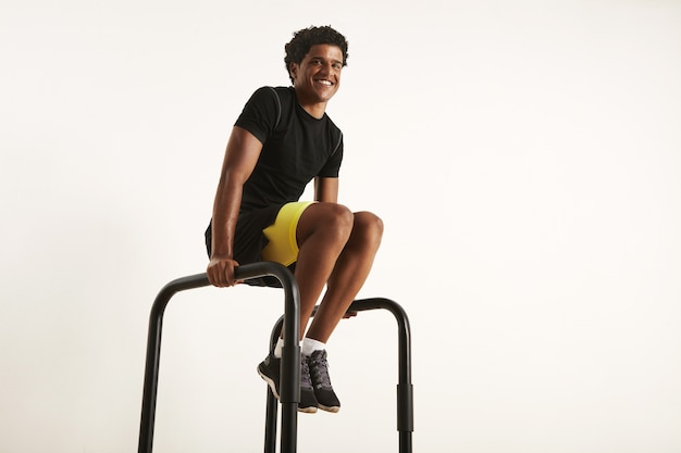 Feliz sonriente hombre afroamericano en equipo de entrenamiento sintético negro haciendo ejercicio en casa en barras paralelas, aislado en blanco