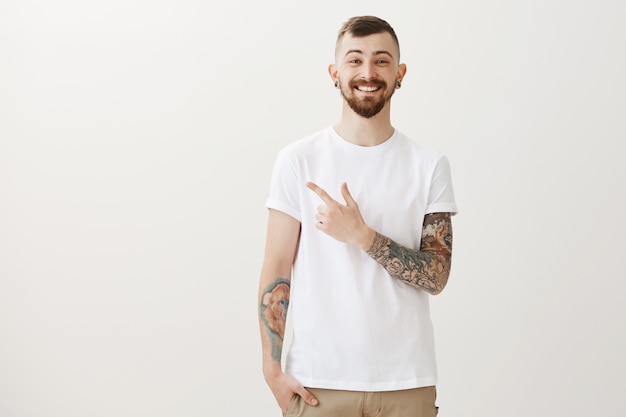 Feliz sonriente hombre barbudo con tatuajes y piercing dedo acusador izquierdo