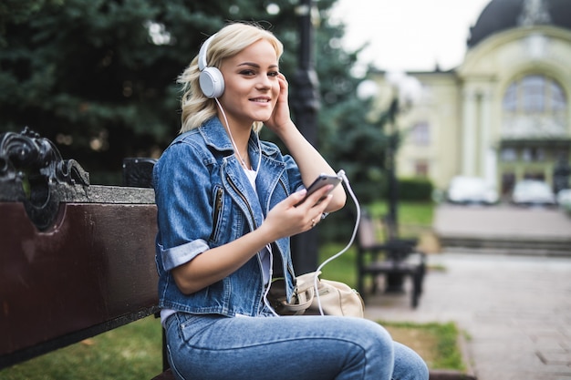 Feliz sonriente joven escuchando música en auriculares y usando el teléfono inteligente mientras está sentado en el banco en la ciudad