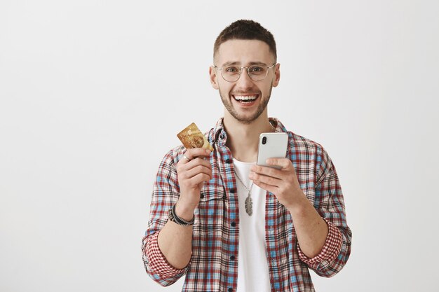 Feliz sonriente joven con gafas posando con su teléfono y tarjeta