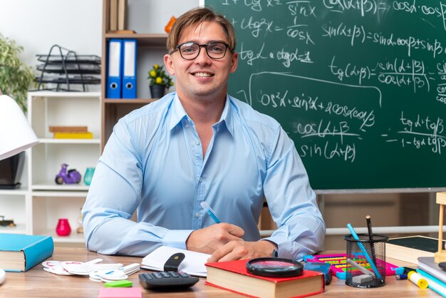 Feliz y sonriente joven profesor con gafas sentado en el escritorio de la escuela con libros y notas delante de la pizarra en el aula