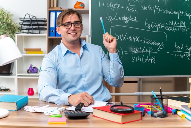 Feliz y sonriente joven profesor sentado en el escritorio de la escuela con libros y notas sosteniendo un lápiz delante de la pizarra en el aula