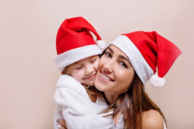 Feliz, sonriente madre e hija divertidas están posando y sosteniendo sus rostros con gorras de Santa. Una joven hermosa con labios brillantes sostiene a una niña de 5 años.