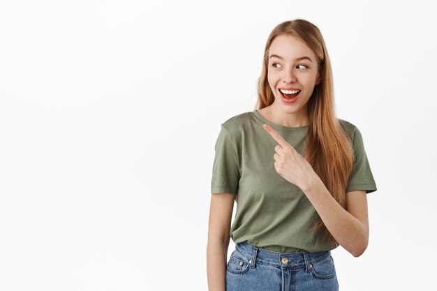 Feliz sonriente modelo femenino apuntando, mirando a la izquierda y riendo sobre pancarta, mostrando algo divertido a un lado, de pie en camiseta contra la pared blanca