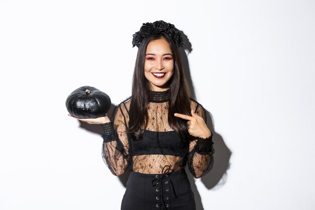 Feliz sonriente mujer asiática disfrutando de la celebración de halloween, vistiendo traje de bruja y señalando con el dedo a la calabaza negra, de pie sobre fondo blanco.