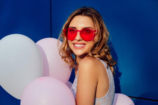 Feliz sonriente mujer joven en gafas de sol con estilo rojo, sosteniendo globos de aire blanco