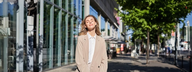 Foto gratuita una feliz y sonriente mujer de negocios cierra los ojos parada afuera y disfruta del cálido sol posando con un traje beige