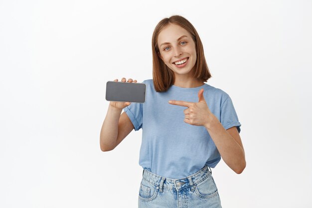 Feliz sonriente mujer rubia presenta la aplicación, apuntando con el dedo a la pantalla horizontal del teléfono inteligente y luciendo satisfecha, recomendando la función del teléfono móvil, tienda de exhibición, pared blanca