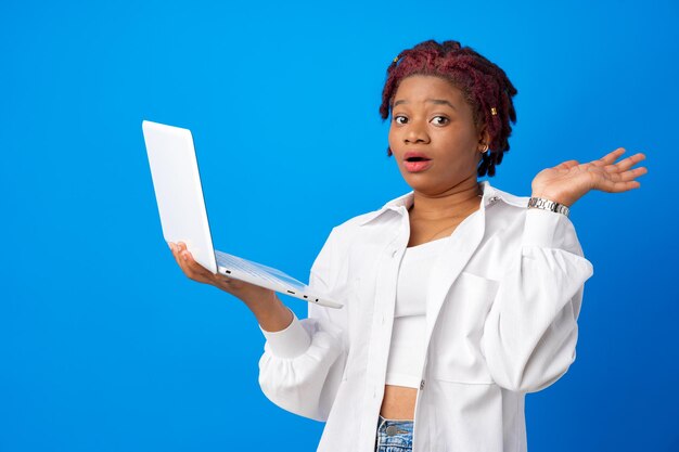 Feliz sorprendida joven afroamericana sosteniendo portátil sobre fondo azul.