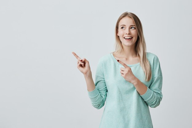 Feliz sorprendida joven rubia sonriendo ampliamente, señalando con el dedo, mostrando algo interesante y emocionante