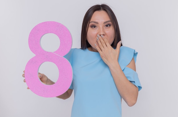 Foto gratuita feliz y sorprendida mujer asiática sosteniendo el número ocho hecho de cartón mirando al frente sonriendo alegremente celebrando el día internacional de la mujer de pie sobre una pared blanca