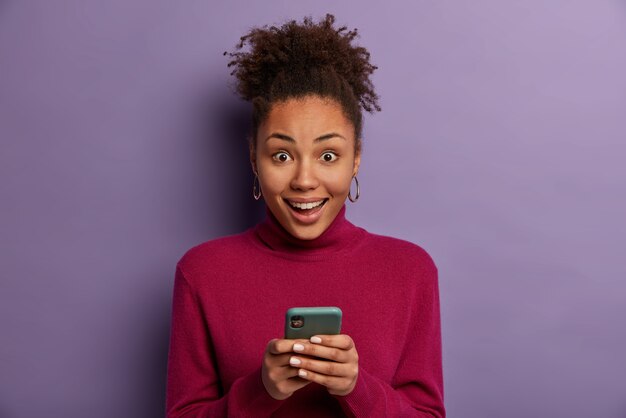 Feliz sorprendida mujer de pelo rizado recibe buenas noticias de un amigo, sostiene el teléfono móvil, envía mensajes de texto, usa un cuello alto informal, aislado en una pared púrpura, descarga la aplicación, navega por Internet