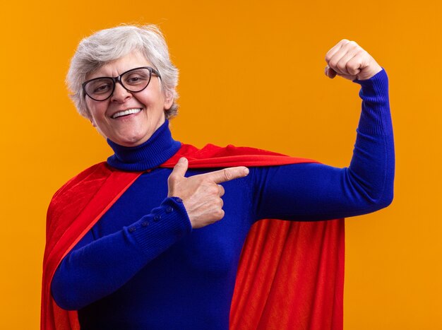 Feliz superhéroe mujer senior con gafas con capa roja posando a la cámara levantando el puño como un ganador mostrando bíceps de pie sobre fondo naranja